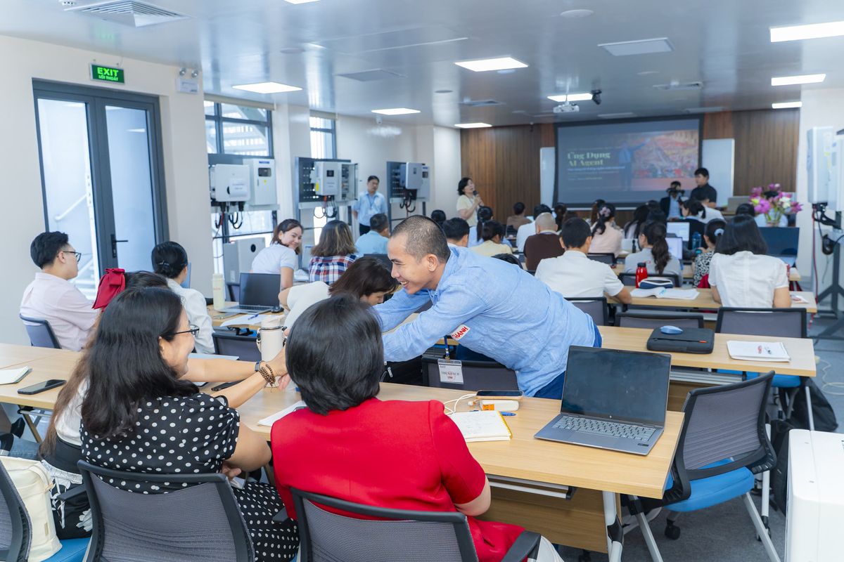 Một người cúi xuống trao đổi, hỗ trợ hai học viên tại bàn; phía trước là màn chiếu Ứng Dụng AI Agent