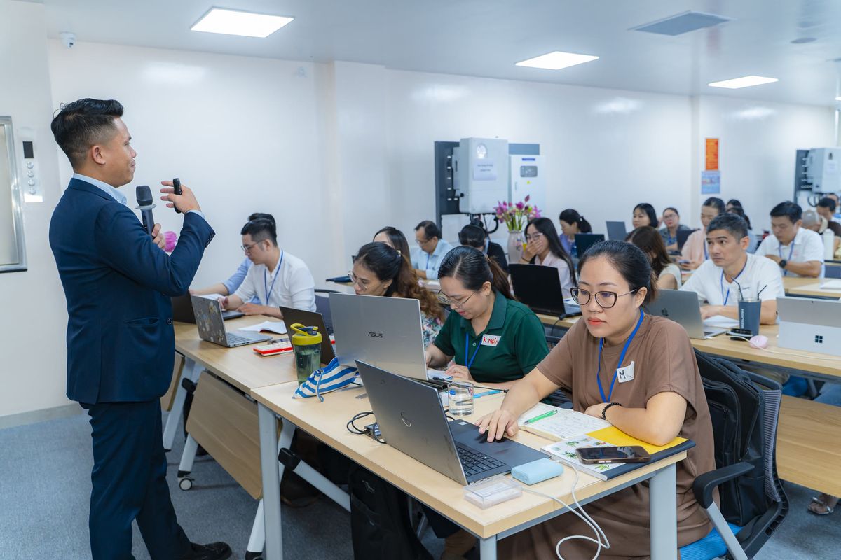 Người hướng dẫn cầm micro đứng cạnh dãy bàn, học viên đang thao tác trên laptop