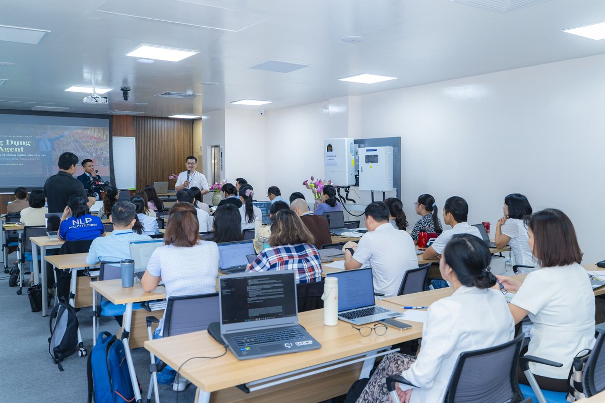 Phòng học nhìn từ cuối lớp, màn chiếu tiêu đề Ứng Dụng AI Agent, học viên mở laptop trước mặt